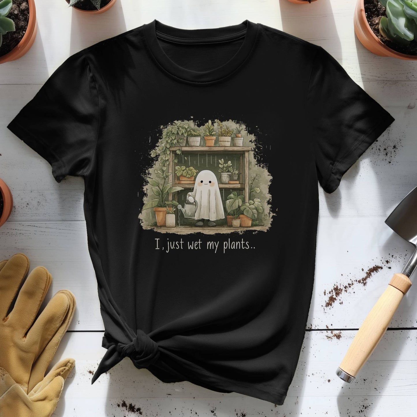Black t-shirt with a ghost and plants design on a white surface with gardening tools and plants.