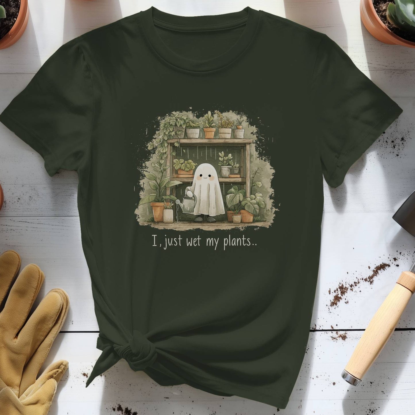 Green t-shirt with a ghost and plants design on a wooden surface with potted plants and gardening tools.