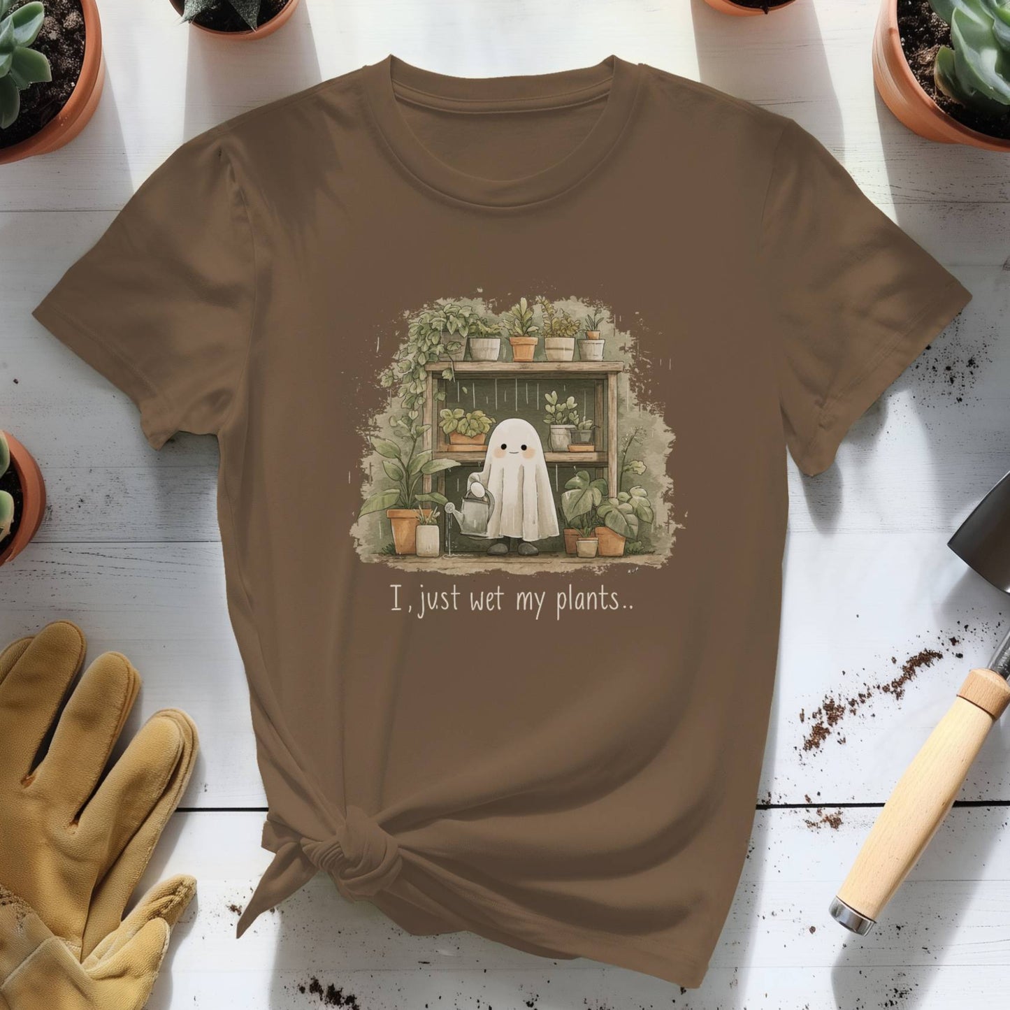 Brown t-shirt with a ghost and plants graphic and text, surrounded by potted plants and gardening tools on a wooden surface.
