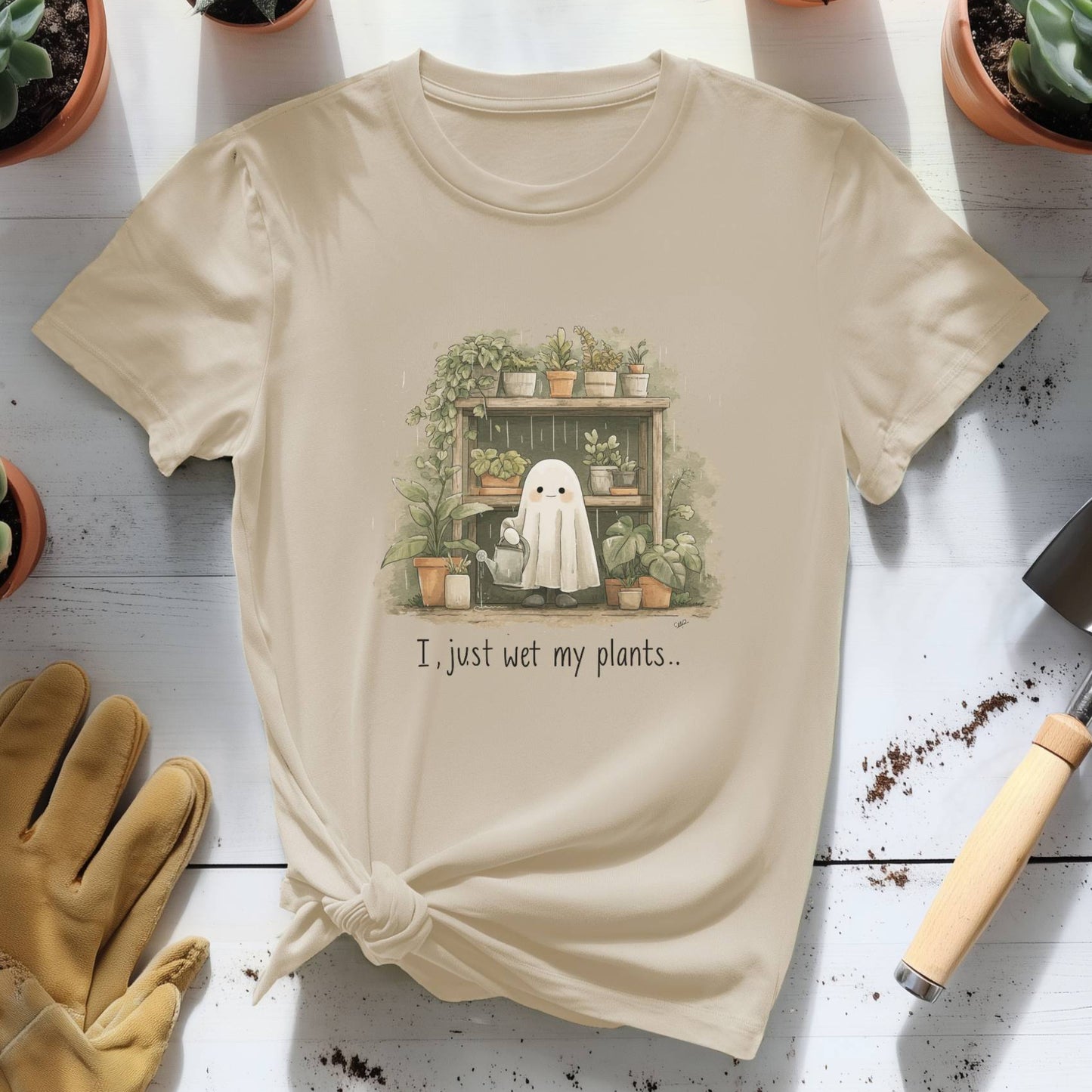 Beige t-shirt with a ghost and plants graphic and text, placed on a wooden surface with gardening tools and plants.