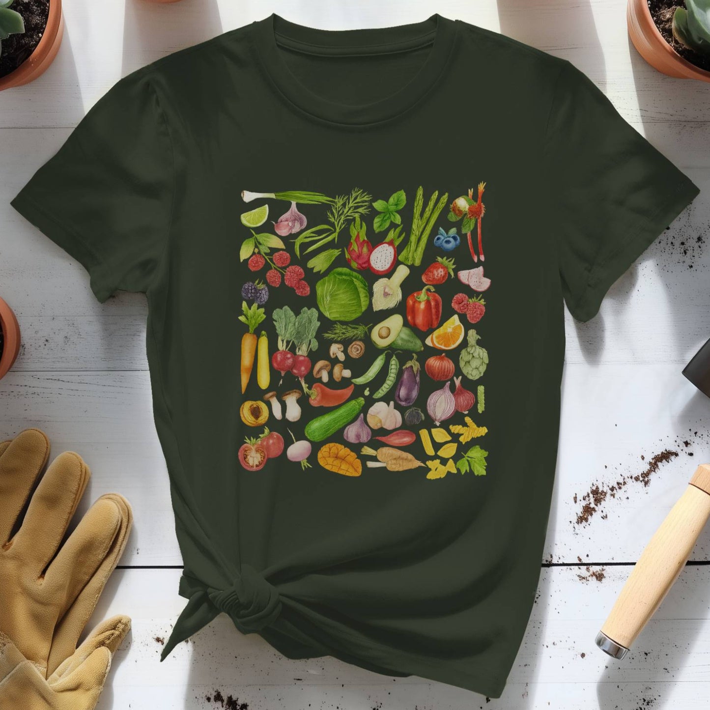 Dark green t-shirt with colorful vegetable pattern on a white wooden surface with gardening tools and plants.