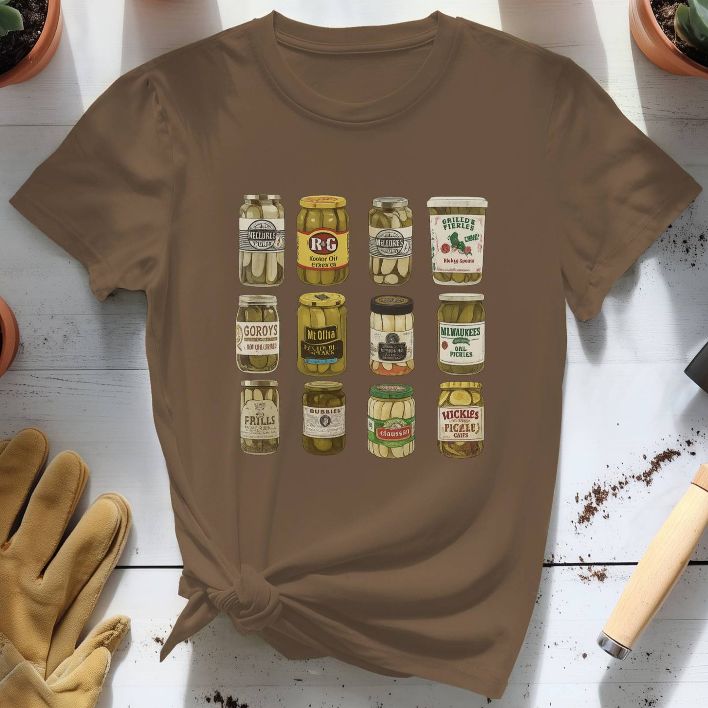 Brown t-shirt with pickle jar graphic on a white wooden surface with plants and gardening tools.