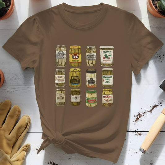 Brown t-shirt with pickle jar graphic on a white wooden surface with plants and gardening tools.